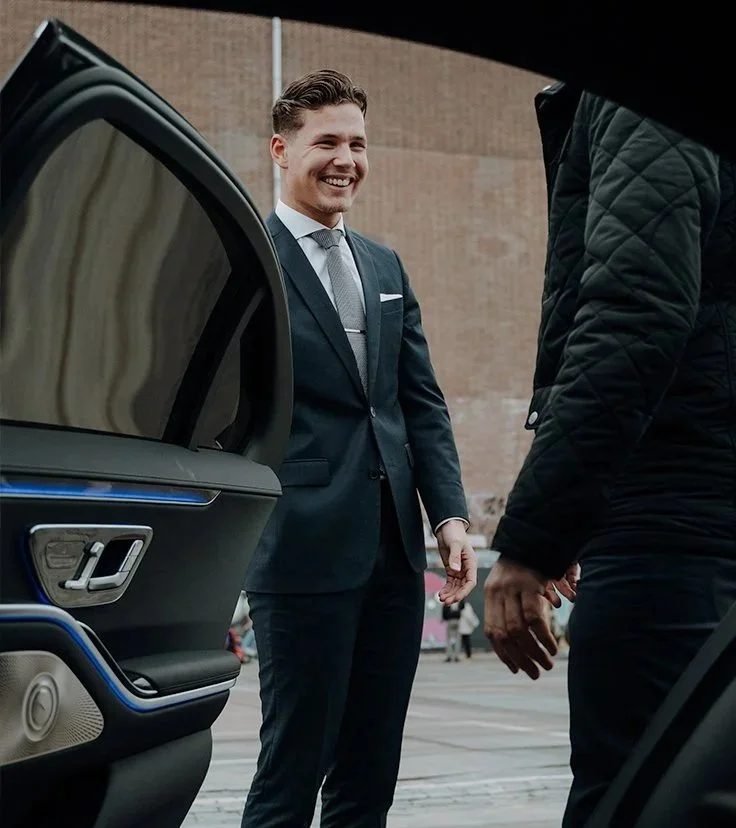 Glimlachende chauffeur in pak bij de open deur van een wagen